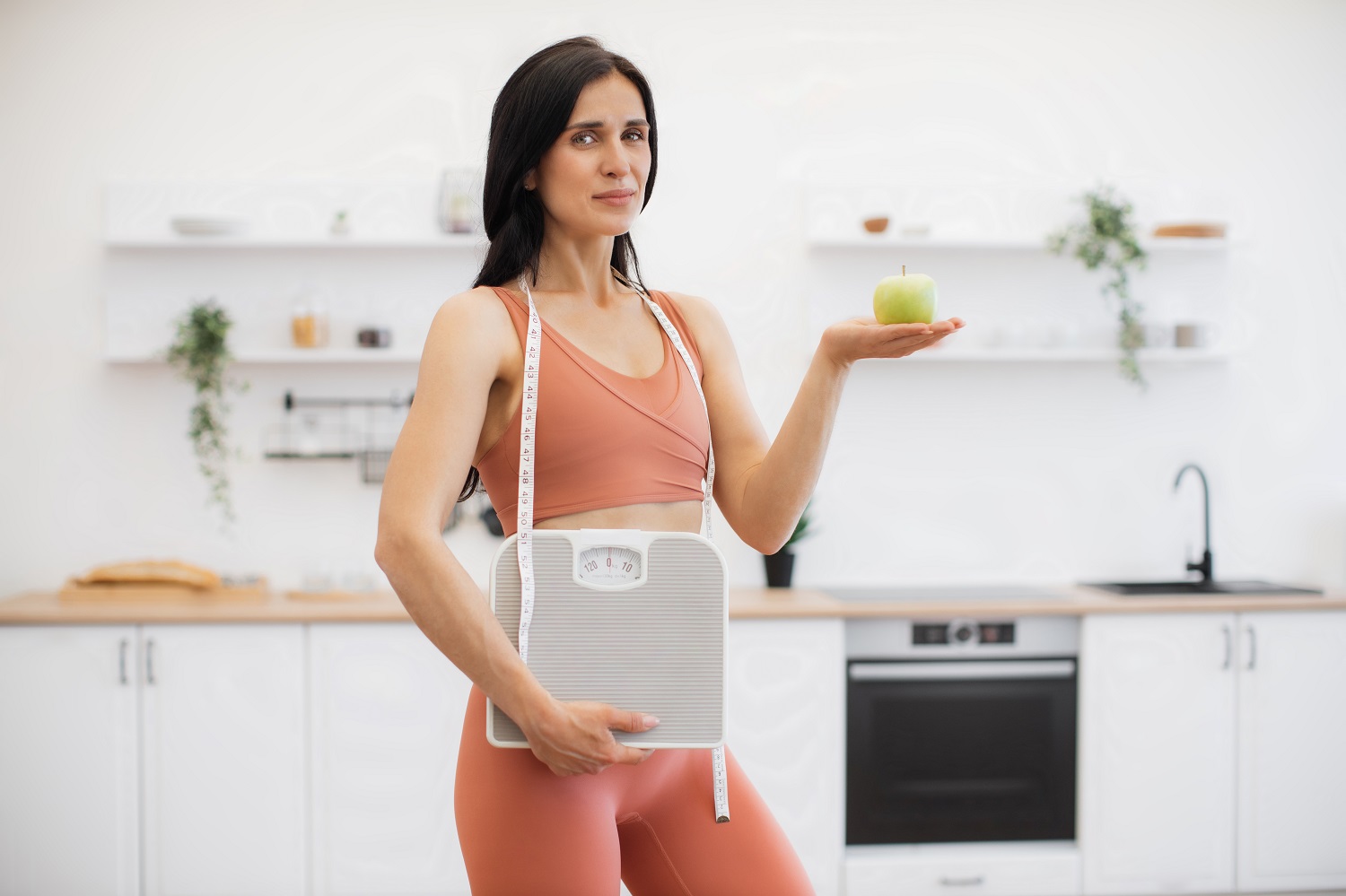 Personal trainer posing with apple and weight scales indoors